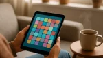 Person on a sofa holding a tablet displaying a colorful calm puzzle game, with a cup of tea and houseplants nearby in a cozy daytime setting.
